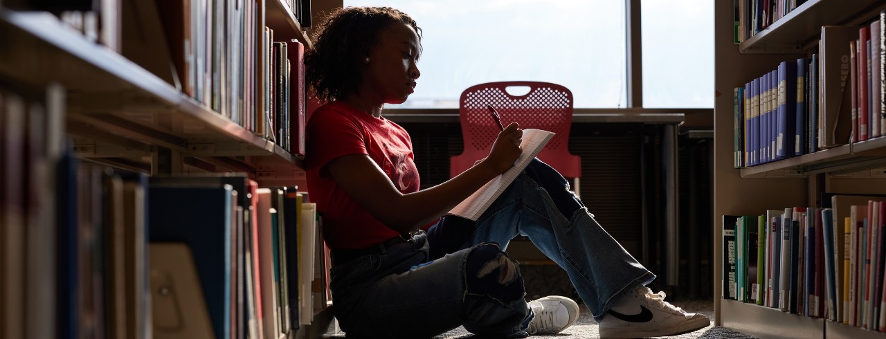 A student writes in a notebook while sitting in the library. 