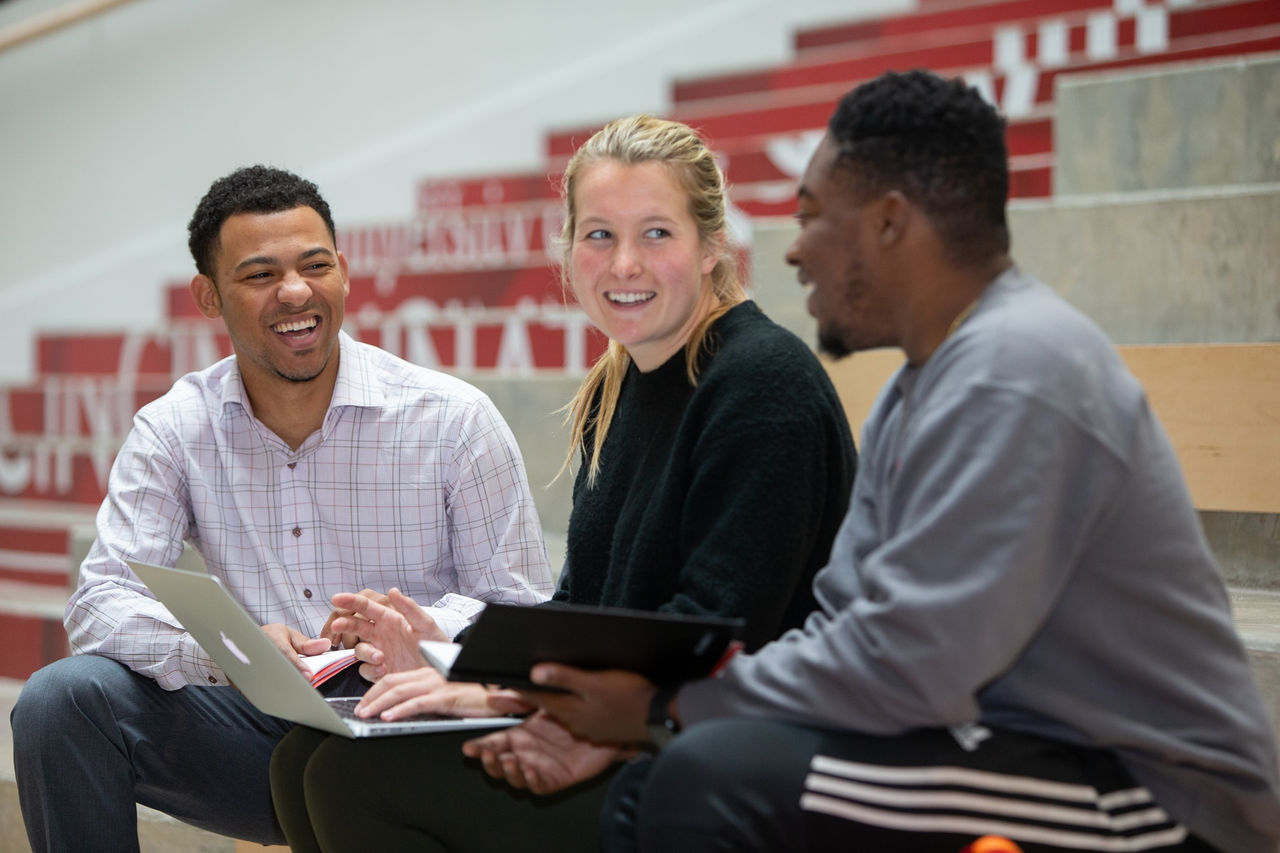 Three people seated on indoor steps in a campus setting, talking together while one uses a laptop and another holds a tablet, with red and white signage visible in the background.
