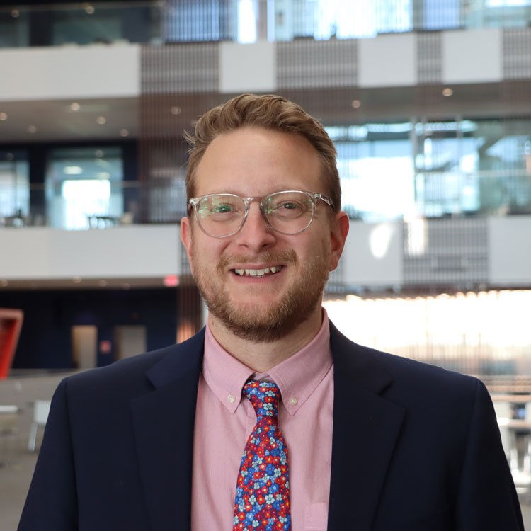 Portrait of a person with short hair and glasses wearing a dark blazer, pink button-down shirt, and patterned tie, standing indoors in a modern, multi-level building with glass and metal architectural details in the background.